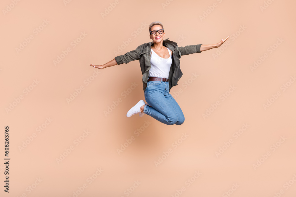 Full length photo of sweet excited lady dressed khaki shirt jumping high arms sides emtpy space isolated beige color background Full length photo of sweet excited lady dressed khaki shirt jumping high arms sides emtpy space isolated beige color background