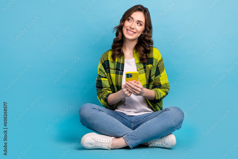 Full length photo of positive person sit floor hold telephone look empty space isolated on blue color background Full length photo of positive person sit floor hold telephone look empty space isolated on blue color background