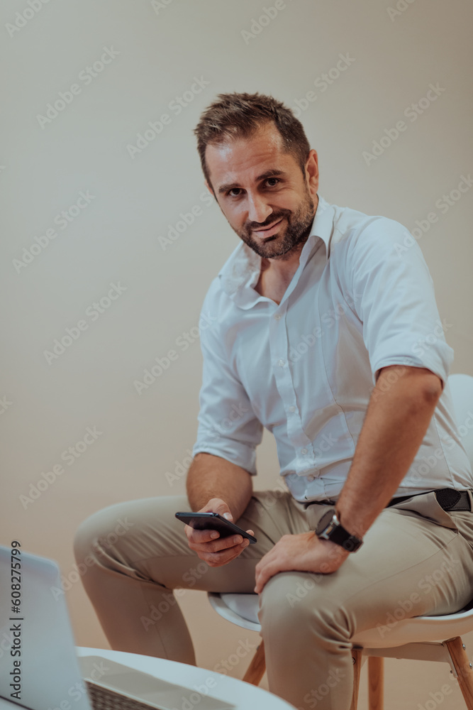 A confident businessman sitting and using laptop and smartphone with a determined expression, while a beige background enhances the professional atmosphere, showcasing his productivity and expertise.