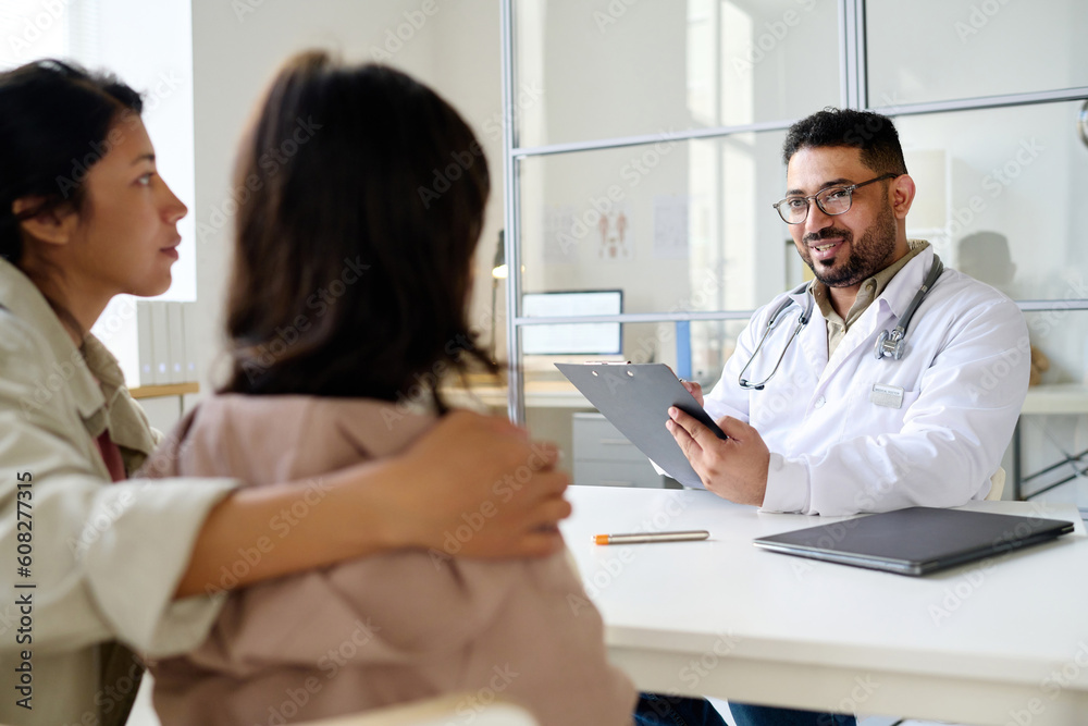 Pediatrician talking to little patient while she sitting opposite him together with her mom during their visit to clinic