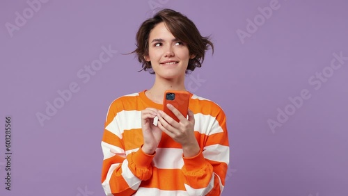 Dreamful happy pensive minded young caucasian woman wear casual clothes hold use mobile cell phone typing browsing chatting send sms isolated on plain pastel light purple background studio portrait