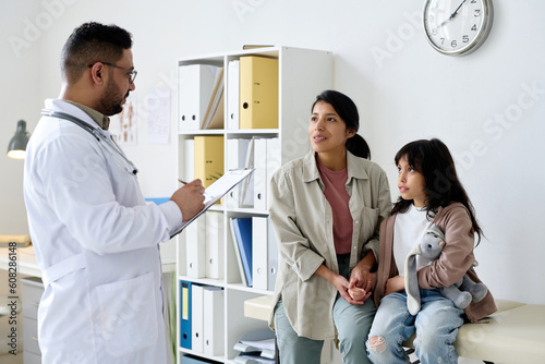 Young mom talking to doctor about the health of her child and doctor making notes in card during their visit in hospital photo