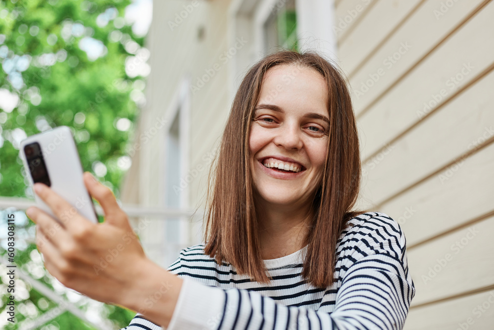 Laughing young woman holding her smartphone in hands while sitting on porch of house using device to send message or browsing internet looking at camera expressing positive emotions. Laughing young woman holding her smartphone in hands while sitting on porch of house using device to send message or browsing internet looking at camera expressing positive emotions.