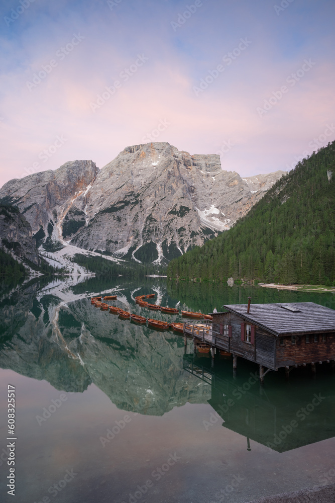 Grüner See in den Bergen mit Booten und einer Holzhütte mit glattem Wasser und Spiegelung vor Sonnenaufgang mit farbigen Wolken.