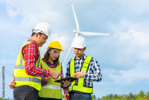 A team of 3 Asian male and female engineers meeting for a project together. point to tablet There's a wind turbine turning into an energy industry business wearing a vest, helmet and radio.