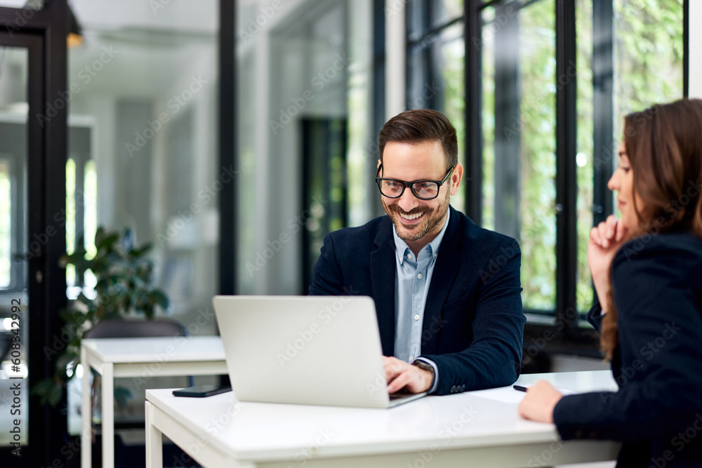 A smiling businessman using a laptop and sitting with a female colleague at the office. A smiling businessman using a laptop and sitting with a female colleague at the office.