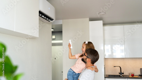 Happy family, father and daughter turn on air conditioner using remote control on background of light interior of apartment.