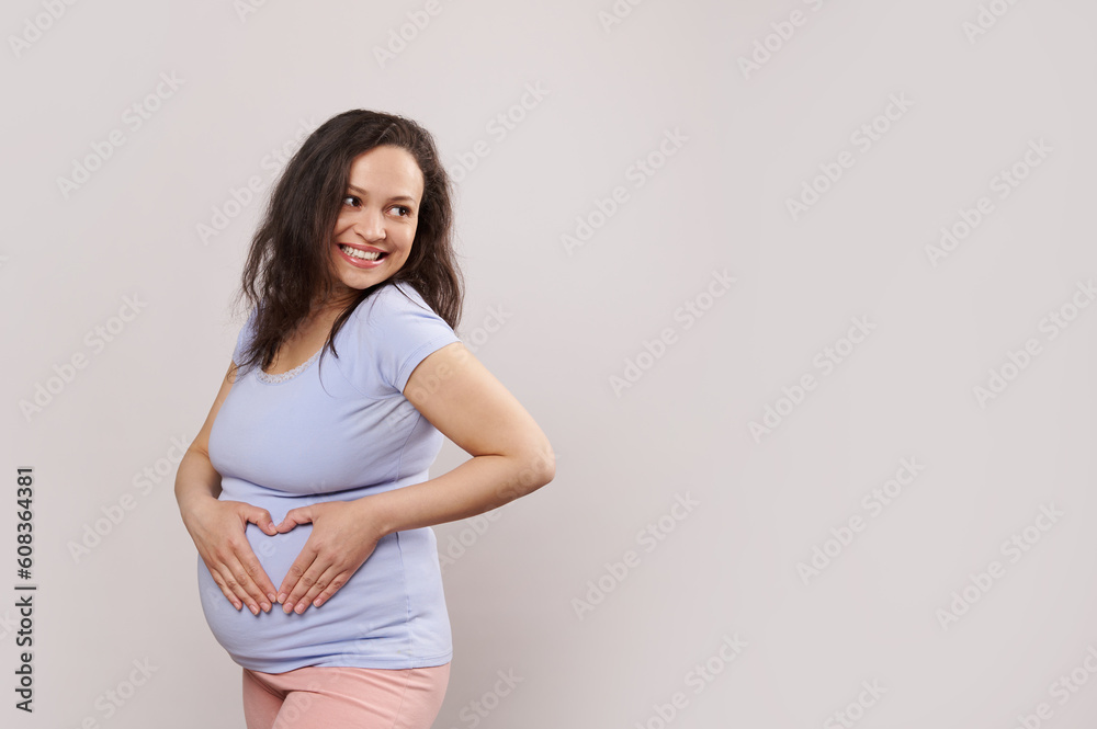 Emotional conceptual portrait delightful happy pregnant woman, expectant mother smiling looking aside, forming heart shape with her hands on belly, isolated on white background. Copy advertising space Emotional conceptual portrait delightful happy pregnant woman, expectant mother smiling looking aside, forming heart shape with her hands on belly, isolated on white background. Copy advertising space