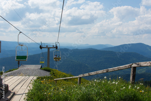 A couple of tourists sitting on a cable car chair high in the mountains on a summer sunny day with a cloudy sky in the background. 