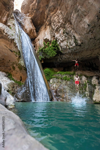 man in red shorts jumping into the azure lake next to a waterfall in a canyon in desert