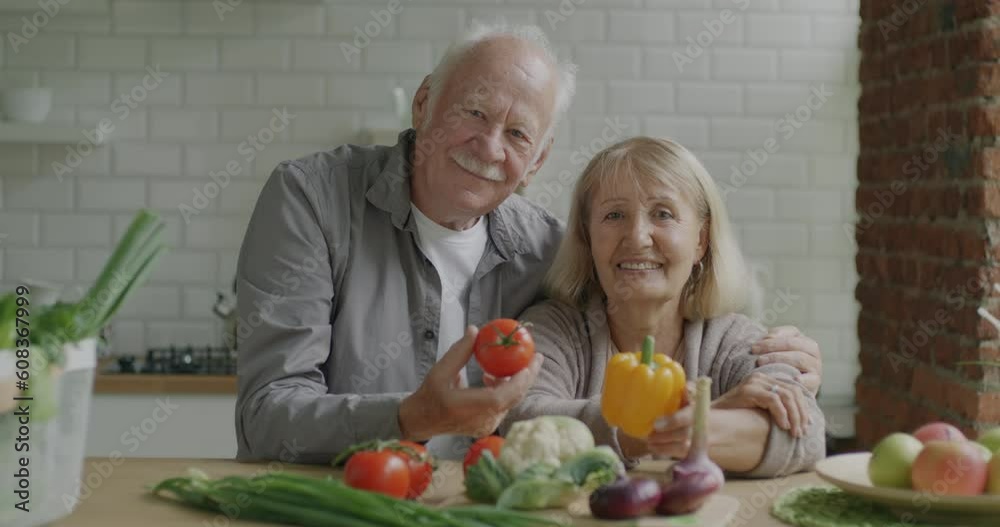 Slow motion portrait of retired man and woman smiling holding fresh vegetables sitting at kitchen table together. Retirement and nutrition concept.