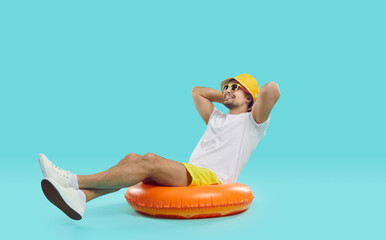 Happy satisfied young man enjoys summer sea vacation sitting on inflatable swimming circle. Joyful guy in summer outfit sits on inflatable circle with his hands behind his head on blue background