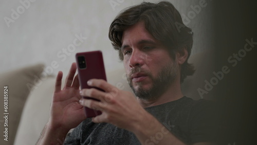 One young man scrolling with cellphone device at home with serious expression. Male person using modern technology sitting on couch