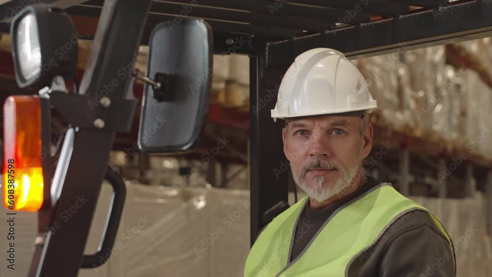 Chest up portrait of mature Caucasian forklift operator looking at camera while sitting at driver seat and speaking on walkie talkie with coworkers during shift at warehouse Chest up portrait of mature Caucasian forklift operator looking at camera while sitting at driver seat and speaking on walkie talkie with coworkers during shift at warehouse