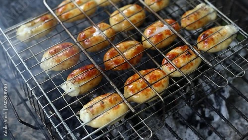 Small grilled sausages on grill with smoke and flame on dark charcoal background outdoor. Baked ruddy sausage grilled on a grill grate. The concept of a picnic in nature. Video fottage in HD 25 FPS photo