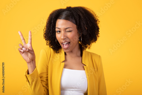 Joyful black curly woman winking and showing peace sign with hand, posing isolated on yellow background, studio shot