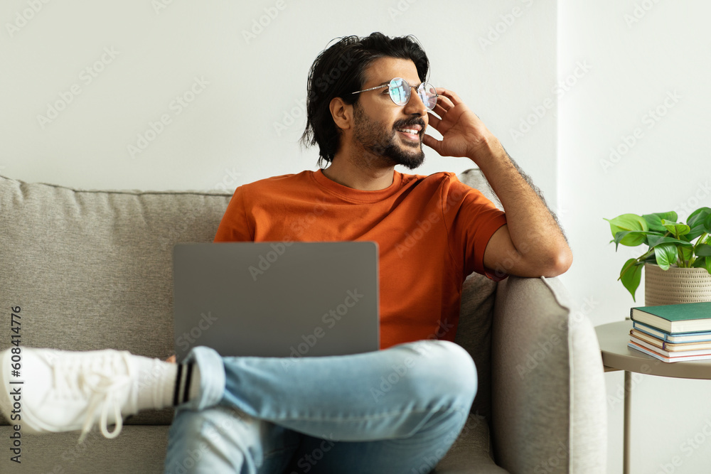 Smiling millennial middle eastern guy in glasses with laptop thinks, looks at empty space, sits on sofa in living room Smiling millennial middle eastern guy in glasses with laptop thinks, looks at empty space, sits on sofa in living room