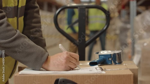 Midsection of unrecognizable warehouse worker using adhesive tape dispenser to pack cardboard box and signing invoice paper, preparing parcel for transportation and dispatching