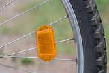 One orange plastic reflector on the gray metal spokes of a bicycle wheel outdoors against a green background.Safety device.