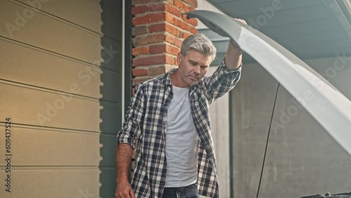 Adult handsome man is looking at his car with the hood open. Mele dressed in shirt and jeans fixing auto engine outside on the background of garage. Person repairs car demages in backyard. photo
