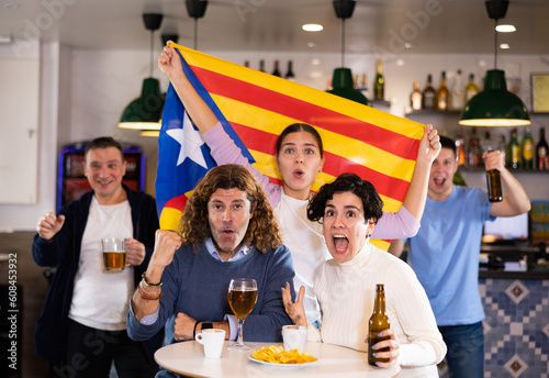 Company of emotional young adult fans supporting favorite team of Estelada with flag while resting in sports bar photo