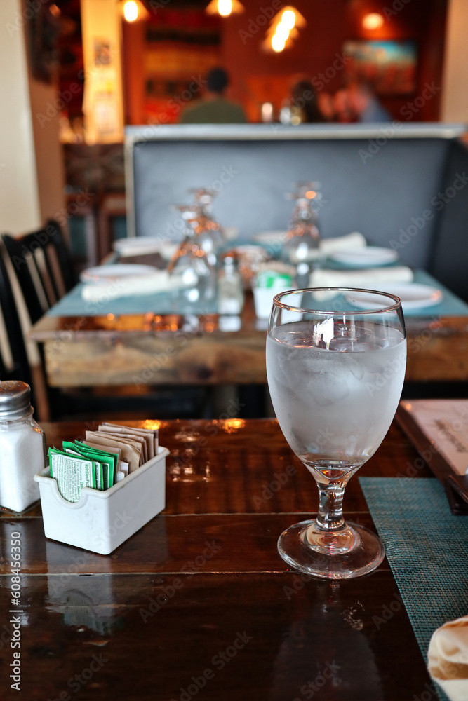 glass of ice water on restaurant table with sugar and tea packets (place settings, chairs, dimly lit eatery)