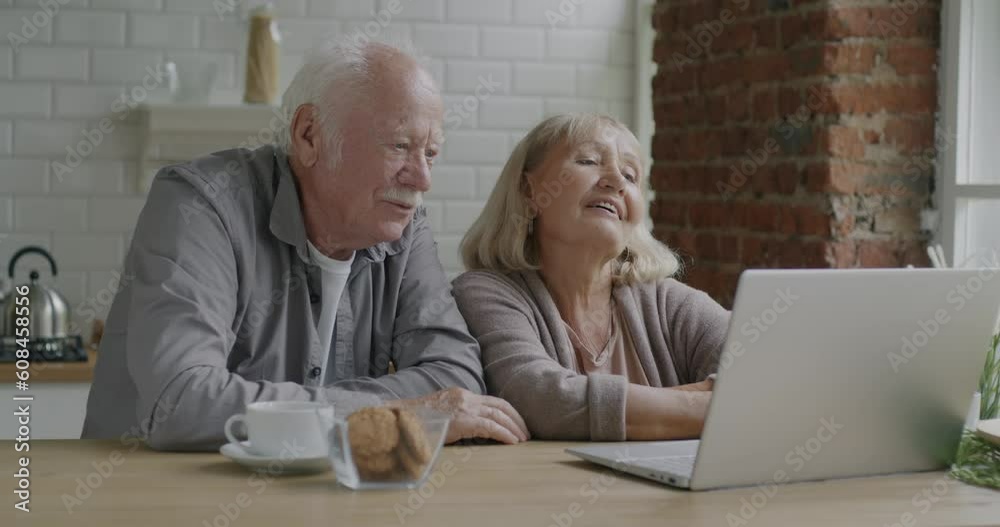 Cheerful senior couple making video call enjoying online communication using laptop computer in kitchen in apartment. Family and modern technology concept. Cheerful senior couple making video call enjoying online communication using laptop computer in kitchen in apartment. Family and modern technology concept.