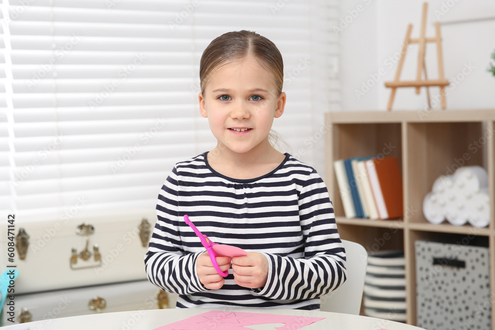 Cute little girl with scissors and pink paper at desk in room. Home workplace Cute little girl with scissors and pink paper at desk in room. Home workplace