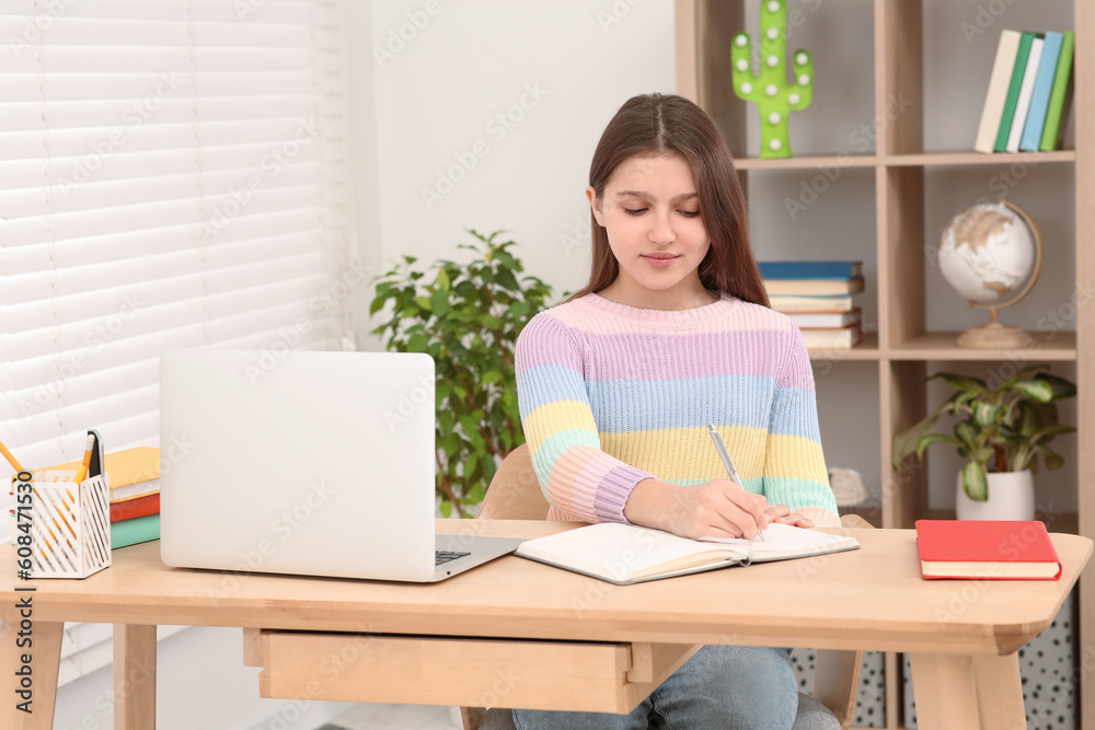 Cute girl writing in notepad near laptop at desk in room. Home workplace