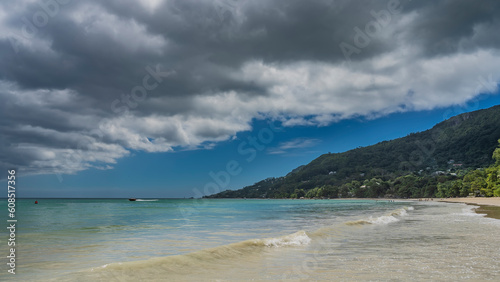 Fototapeta Naklejka Na Ścianę i Meble -  The waves of the turquoise ocean roll to the shore and spread over the sandy beach. Boats are visible in the distance. A green hill against a  blue sky and clouds. Seychelles. Mahe. Beau Vallon