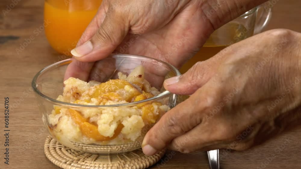 Male senior hands putting a transparent glass bowl of delicious homemade peach cobbler on bamboo mat with a glass of orange juice on wooden table. Male senior hands putting a transparent glass bowl of delicious homemade peach cobbler on bamboo mat with a glass of orange juice on wooden table.