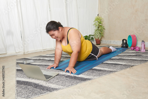 sporty plus size woman in sportswear working out at home, doing plank on yoga mat in front of Video lesson, repeating instructions by professional fitness trainer watching online video tutorial.