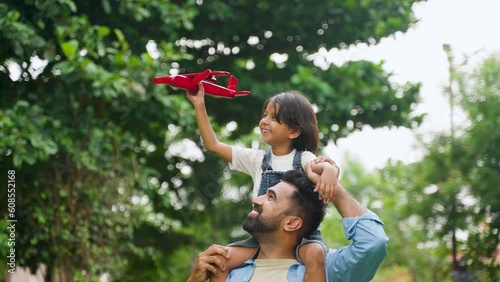 Happy smiling indian girl kid playing with airplane toy by sitting on father shoulder at park - concept of freedom, family support and togetherness