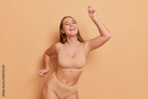 Pretty half-naked woman wearing beige sport bra and panties dances on peach-colour background with her hands up, body comfort concept, copy space