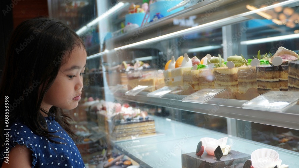 Asian little girl looking at cakes and sweets on display in a bakery shop.