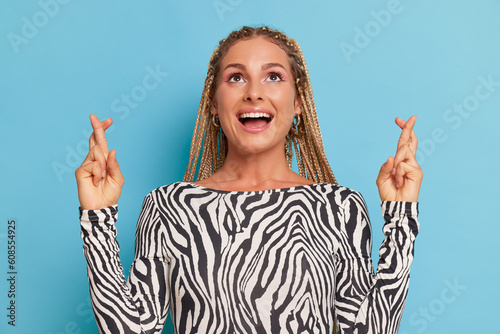 Pretty woman with fair dreadlocks smiles and looks up, poses on blue studio background dressed in black and white sebra style top, lucky time concept, copy space photo
