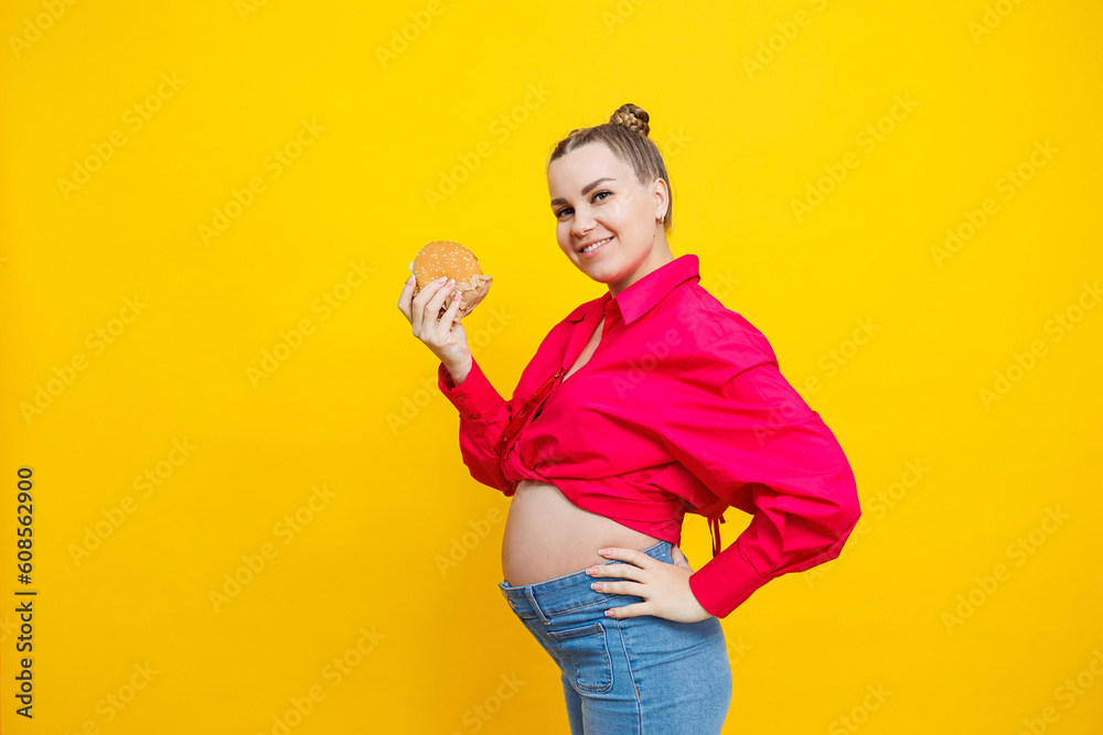 Funny pregnant young woman eating burger. Pregnant woman holding tasty sandwich while standing isolated on yellow studio wall. Junk food concept. Fast food during pregnancy. Funny pregnant young woman eating burger. Pregnant woman holding tasty sandwich while standing isolated on yellow studio wall. Junk food concept. Fast food during pregnancy.