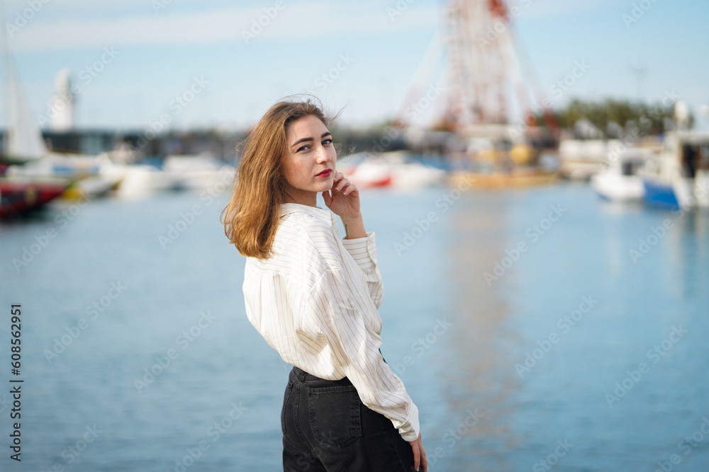 Beautiful girl standing near sea. Seaport or berth in background. Young woman Wearing casual Outfit posing near water in sunny day. People lifestyle and rest concept. Blurred background