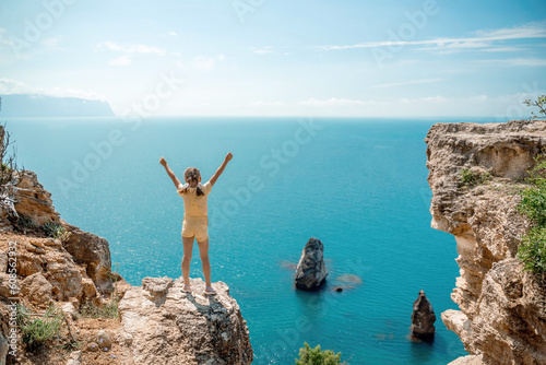 Happy girl stands on a rock high above the sea, wearing a yellow jumpsuit and sporting braided hair, depicting the idea of a summer vacation by the sea.
