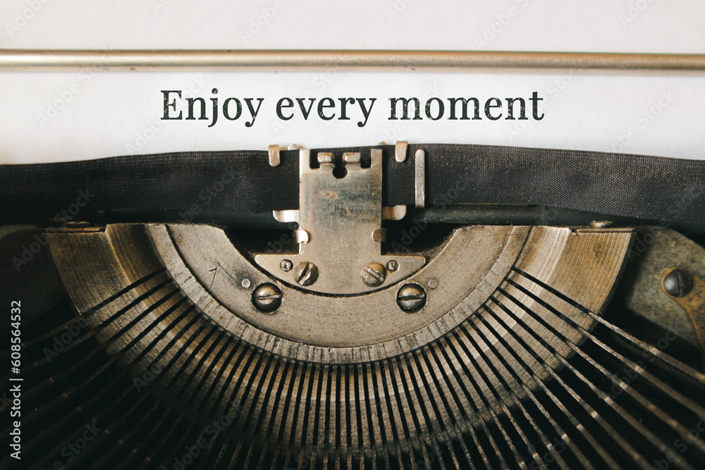 "Enjoy every moment" printed on a sheet of vintage typewriter. - obrazy, fototapety, plakaty 