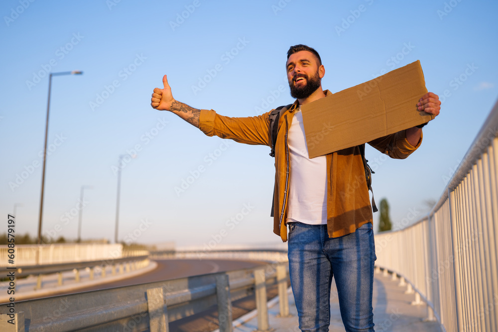 Man is hitchhiking on roadside trying to stop car. He is holding blank cardboard for your text.