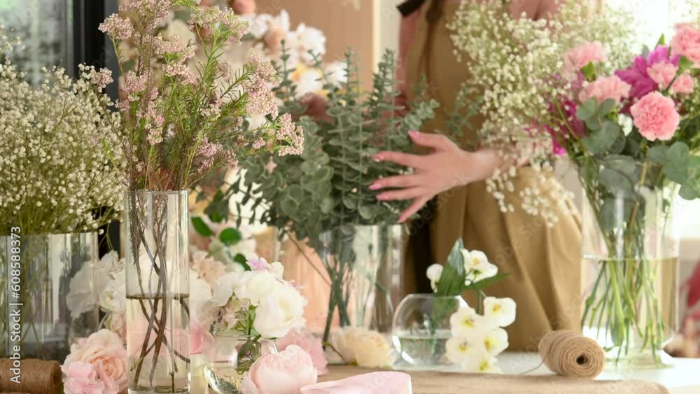 Positive young female florist in green apron standing at table and cutting flowers in floral shop. Woman florist makes a bouquet at home in the decoration studio. Flowers and vases close-up. Positive young female florist in green apron standing at table and cutting flowers in floral shop. Woman florist makes a bouquet at home in the decoration studio. Flowers and vases close-up.