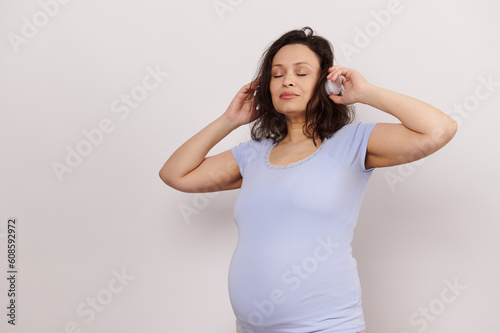 Multi ethnic brunette  happy adult pregnant woman listening to soothing music on wireless headphones  posing with her eyes closed over white isolated background. Happy pregnancy. Maternity concept