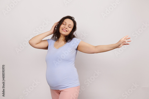 Attractive beautiful pregnant woman expecting baby, listening to music on headphones, enjoying wonderful moments of her pregnancy and maternity lifestyle, posing with eyes closed over white background