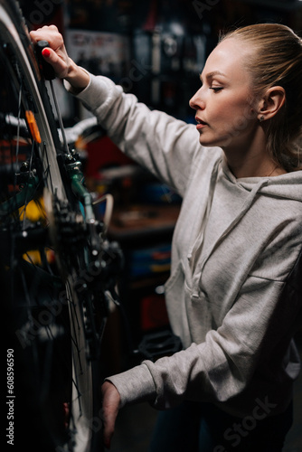 Vertical side view of focused cycling mechanic female repairing and fixing mountain bicycle standing on bike rack in repair workshop with dark interior. Concept of professional bicycle maintenance.