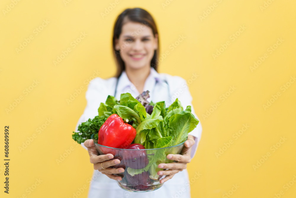 Happy cheerful female doctor or nutritionist showing a vegetables in transparent bowl to camera close up, female doctor holding vegetable basket portrait on yellow. Happy cheerful female doctor or nutritionist showing a vegetables in transparent bowl to camera close up, female doctor holding vegetable basket portrait on yellow.