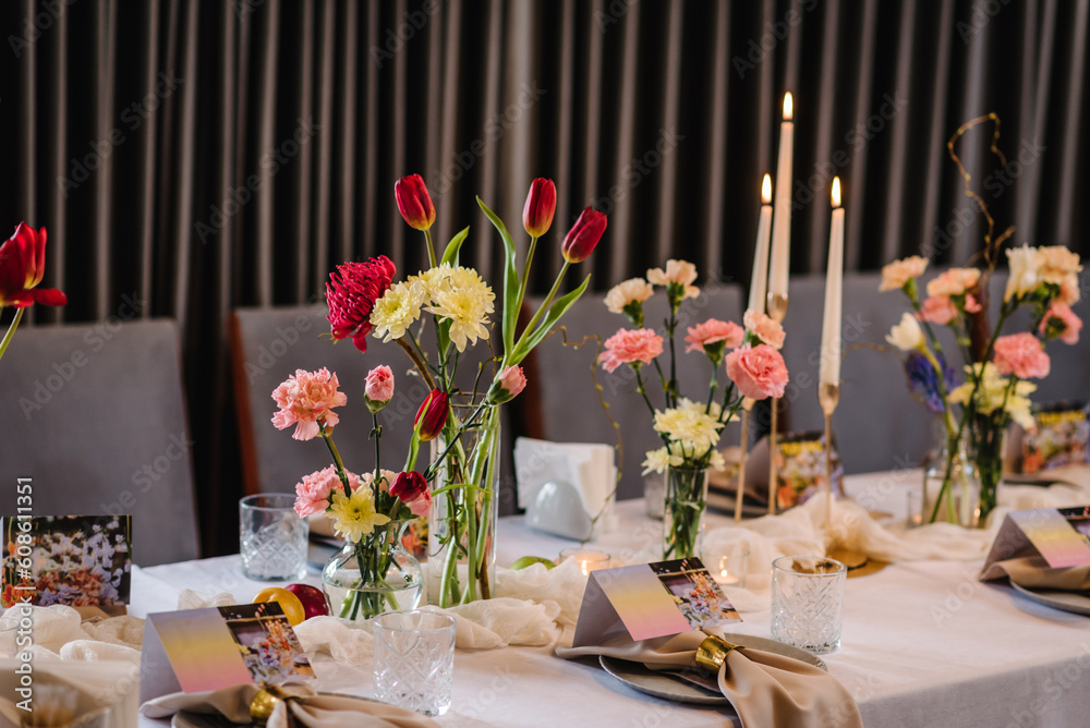 Festive table decorated with composition of red, pink flowers and greenery, candles in hall. Table newlyweds and guest in banquet area on wedding party. Luxurious celebration of event in restaurant.