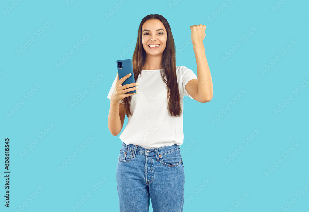 Portrait of happy joyful beautiful young brunette woman in white T shirt and blue jeans standing isolated on blue background, holding mobile phone, doing yes success fist gesture and smiling Portrait of happy joyful beautiful young brunette woman in white T shirt and blue jeans standing isolated on blue background, holding mobile phone, doing yes success fist gesture and smiling