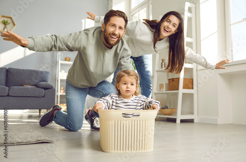 Dad, mom and toddler baby having fun playing at home. Cheerful parents playing together flying like airplane, spreading his arms to sides, cute kid sitting in plasatic box in living room at home photo