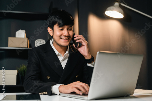 Young asian businessman working with smartphone in the dark office at night. Attractive handsome Indian man work hard overtime in home with floor lamp ambient warm light late at night